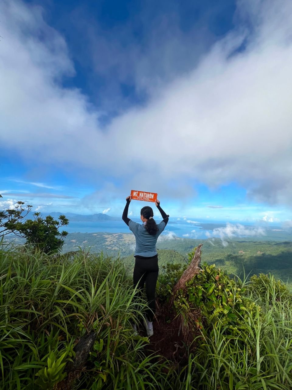 Hiker holding a Mt. Naturon sign above the mountain and coastal views